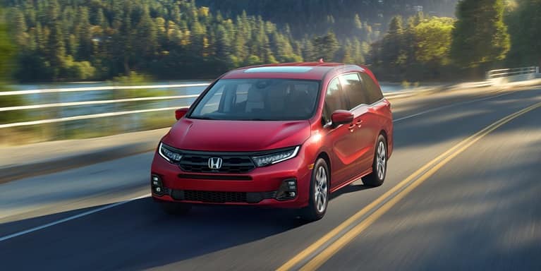 2027 Honda Odyssey Elite in Radiant Red Metallic II front driving on a scenic road near a lake and forest (Minivan).