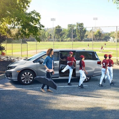 2027 Honda Odyssey Elite in Solar Silver Metallic parked at a baseball field with children, showcasing a spacious minivan for active families (Minivan).